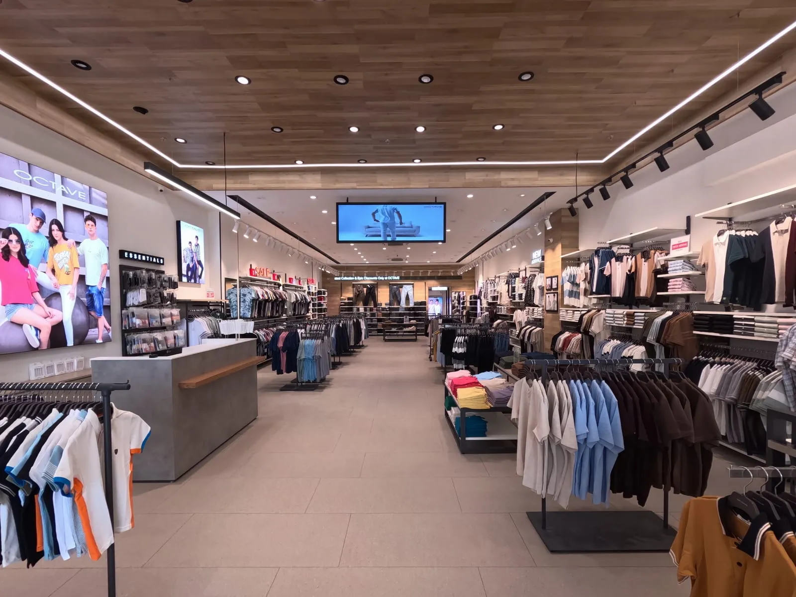 Inside an OCTAVE outlet: menswear on rail and shelf displays either side of a central aisle, under track lighting and a wood-panelled ceiling, with an overhead LED screen and a digital ticker running above the rear wall.